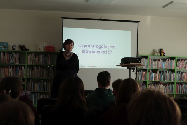 Na zdjęciu znajduje się kobieta, która stoi obok ekranu, na którym wyświetlana jest prezentacja o słowiańskości przez stojący przed nim rzutnik. Przed kobietą siedzi młodzież. W tle znajdują się półki z książkami.