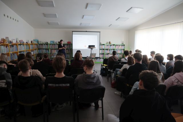 Na zdjęciu znajduje się siedząca młodzież skierowana w stronę kobiety, która przedstawia prezentację na temat Słowian. W tle znajdują się półki z książkami oraz okna po prawej stronie.