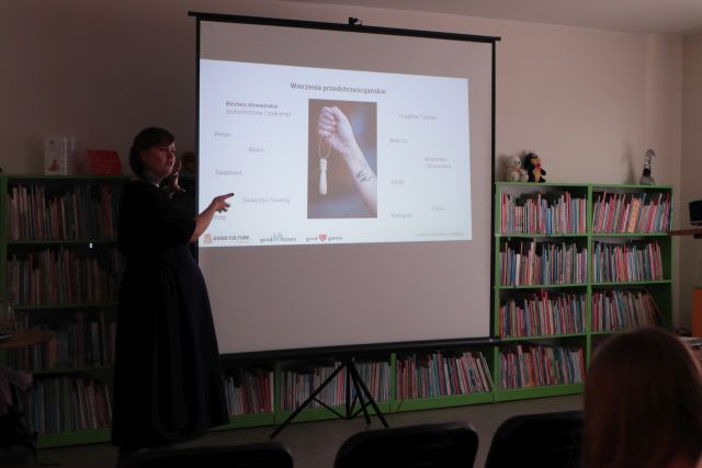 Na zdjęciu znajduje się kobieta, która stoi obok ekranu, na którym wyświetlana jest prezentacja o wierzeniach przedcheścijańskich. W tle znajdują się półki z książkami.