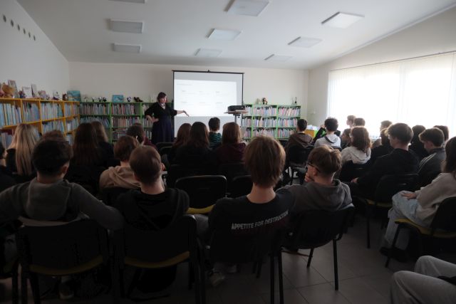 Na zdjęciu znajduje się młodzież siedząca na wprost kobiety stojącej obok rzutnika. W tle znajdują się półki z książkami.