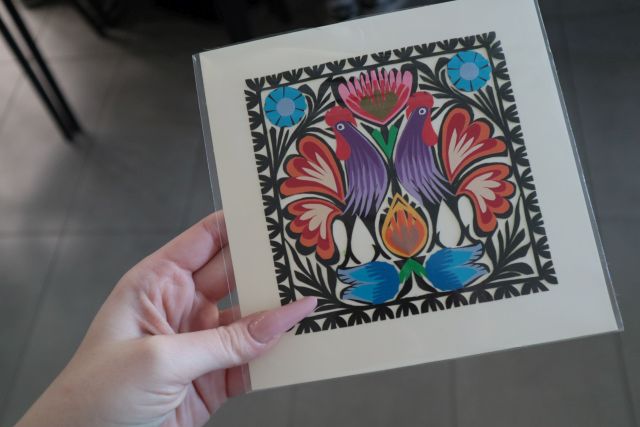 Na zdjęciu znajduje się kobieca dłoń trzymająca ludową wycinankę przedstawiająca dwa koguty. 