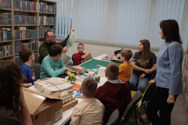 Na zdjęciu znajdują się chłopcy oraz trójka rodziców przy długim stole, na którym znajdują się materiały do tworzenia makiety, jeden siedzący mężczyzna rozwija mapę. Za stołem stoi kobieta. W tle znajdują się okna i półki z książkami po lewej stronie.