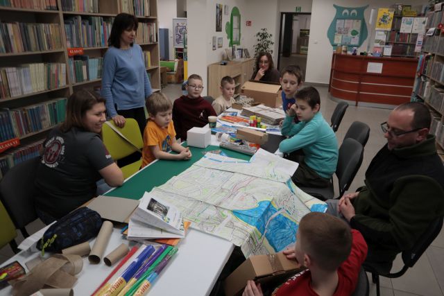 Na zdjęciu znajduje się sześciu chłopców oraz trzech dorosłych przy stole. Przed chłopcem i mężczyzną obok leży rozłożona mapa, za stołem na przeciwko stoi kobieta. Na stole leżą materiały do stworzenia makiety miasta kleje, kartony, rolki po papierze. W tle znajdują się półki z książkami. 