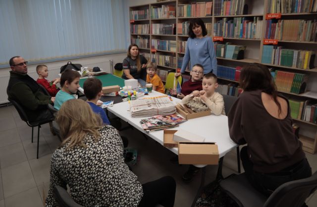 Na zdjęciu znajduje się czwórka dorosłych i sześciu chłopców przy stole, na którym znajdują się gazety, kartki, kartony, kleje do stworzenia makiety miasta. Za stołem po prawej stronie stoi kobieta. W tle znajduja się okna oraz półki z książkami.
