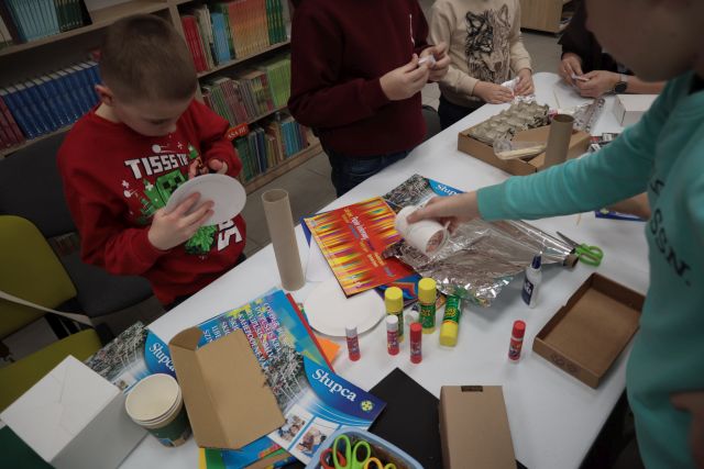 Na zdjęciu znajduje się chłopiec sięgający rolke ze stołu, na którym leżą materiały do stworzenia makiety. Na przeciwko siedzi dwójka chłopćow, która wycina i klei elementy do makiety. W tle znajdują się półki z książkami.