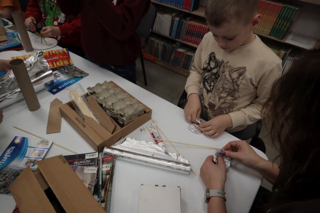 Na zdjęciu znajduje się chłopiec, który klei fragment do makiety, na stole przed którym stoi leżą kartony, folia aluminiowa, pomaga mu kobieta siedząca obok niego. W tle znajdują się półki z książkami. 