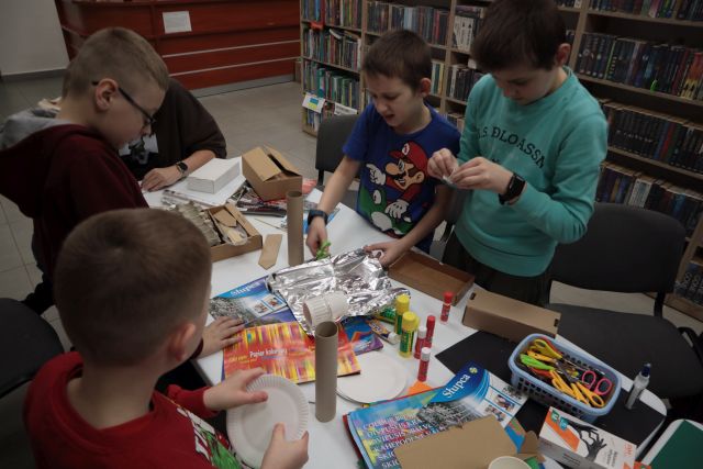 Na zdjęciu znajduje się czterech chłopców oraz jedna pochylona w dół kobieta przy stole i wykonują oni elementy do makiety z materiałów znajdujących się na stole, kartek, foli aluminiowej, kleju kolorowych kartek. W tle znajdują się półki z książkami.