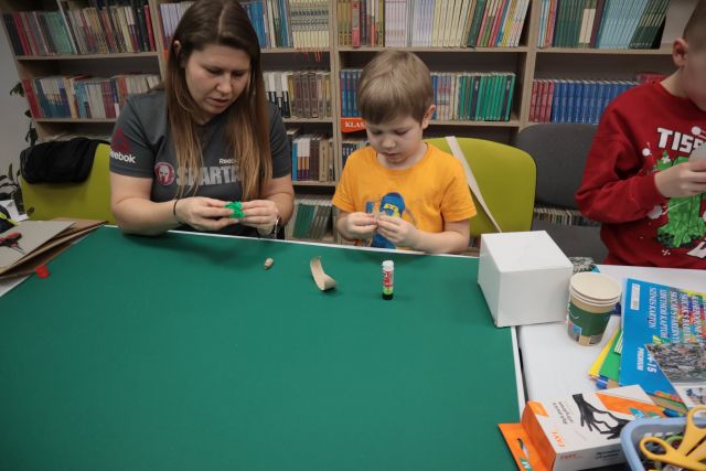 Na zdjęciu znajduje się chłopiec z mamą przed nimi leży zielony brystyl, do którego przyklejają część makiety. Obok chłopca znajduje się chłopiec w czerwonej bluzce, który trzyma w rękach kawałek kartonika, a przed nim znajdują się kolorowe kartki, nożyczki. W tle znajdują się półki z książkami.