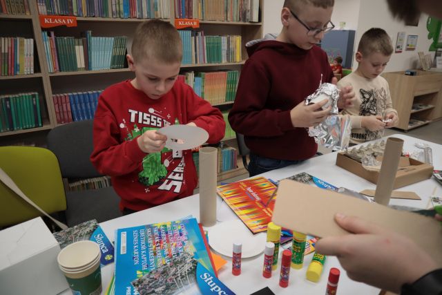 Na zdjęciu znajduje się trójka chłopców, którzy wycinają i kleją budynki z kartoników, papieru i foli aluminiowej z leżących przed nimi materiałów. Za nimi znajdują się półki z książkami, a przed uchwycona jest ręka chłopca, który wycina coś z kartonu.