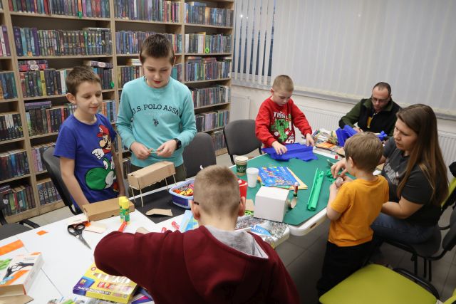 Na zdjęciu znajduje się czterech chłopców oraz kobieta z mężczyzną przy jednym długim stole, na którym znajdują się materiały do stworzenia makiety, uczestnicy z ich pomocą tworzą budowle. W tle znajdują się okna oraz półki z książkami.