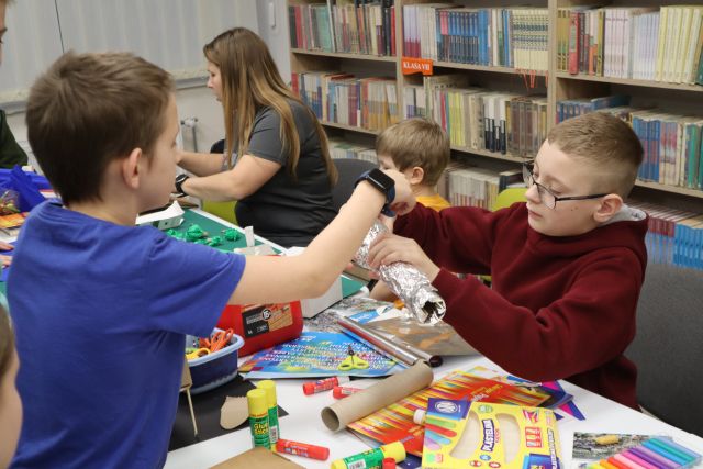 Na zdjęciu znajduje się trójka chłopców oraz kobieta. Jeden chłopiec pomaga drugiemu chłopcu po drugiej stronie stołu. Na stole znajdują się bloki kolorowe, plastelina, kleje, nożyczki, rolki. W tle znajdują się półki z książkami.
