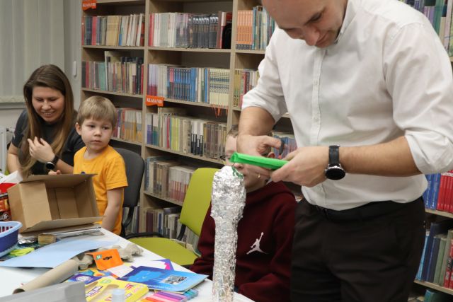 Na zdjęciu znajduje się mężczyzna ucinający bibułę przed nim na stole leży makiety z wieżą ciśnień ze sreberka, obok niego siedzi chłopiec. W tle przy stole znajduje się chłopiec z mamą. W tle znajdują się kolorowe kartki i kartony. W tle znajdują się półki z książkami.