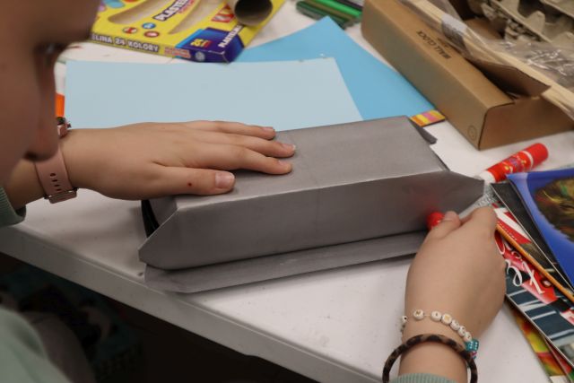 Na zdjęciu znajdują się ręce dziewczynki oraz kawałek jej twarzy, dziewczynka obkleja karton szarą kartką, obok jej ręki znajdują się kolorowe kartki, na przeciwko kartony oraz drewniane patyczki.