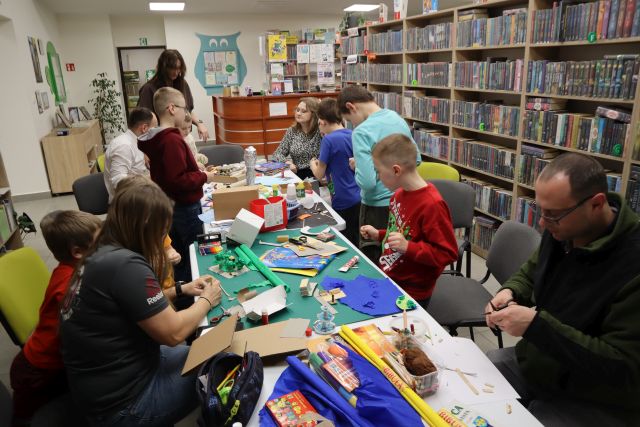 Na zdjęciu znajduje się sześciu chłopców, trzy kobiety oraz dwóch mężczyzn przy stole. Część stoi, część siedzi, na stole znajdują się kolorowe kartki, kartony, kleje i inne materiały potrzebne do stworzenia makiety, którą wykonują uczestnicy warsztatów. W tle znajdują się półki z książkami.