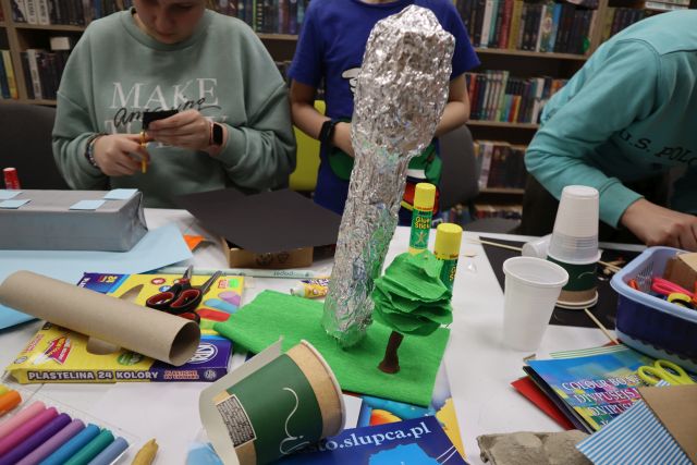 Na zdjęciu znajduje się fragment makiety przedstawiający wieże ciśnień z foli aluminiowej oraz papierowe drzewo obok. W tle znajduje się dwóch chłopców oraz dziewczyna, którzy kleją i wycinają kolejne elementy. Na stole znajdują się papierowe kubeczki, kleje, kolorowe kartki, a w tle półki z książkami.