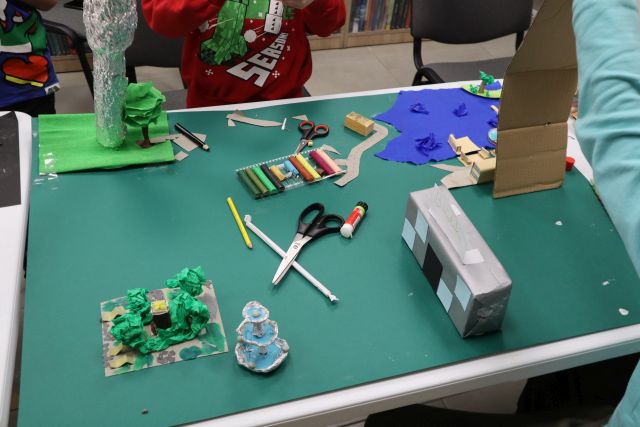 Na zdjęciu znajduje się niedokończona makieta przestawiająca miasto Słupca z Biblioteką, fontanną, wieżą ciśnień, jeziorem. Na makiecie znajdują się również nożyczki, kleje, plastelina. W tle przy stole znajduje się chłopiec.