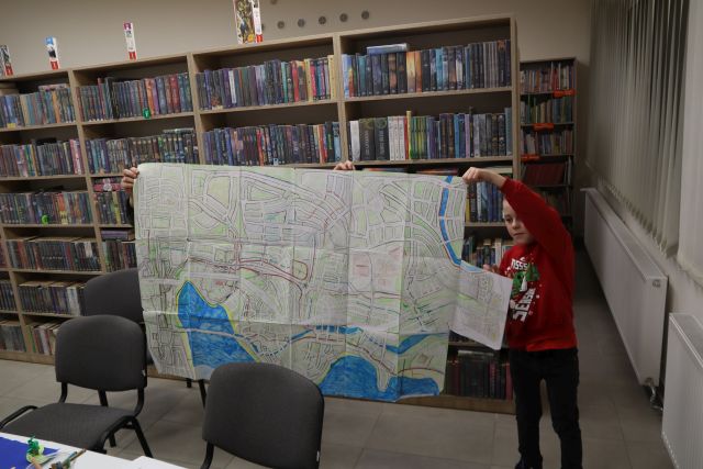 Na zdjęciu znajduje się dwóch chłopców, którzy trzymają rozłożoną dużą mapę, przed nimi stoją dwa krzesła. a za nimi znajdują się półki z książkami.