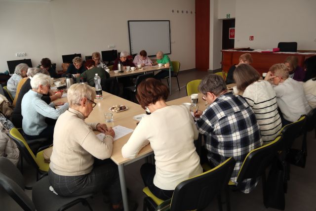 około 20 seniorek siedzi przy stołach ustawionych w literę u i pisze na kartkach, w tle biurka z komputerami