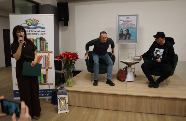 Na zdjęciu znajduje się dwóch mężczyzn siedzących na scenie, za nimi znajduje się plakat promujący spotkanie. Po lewej stronie przy scenie stoi kobieta z mikrofonem zapowiadająca spotkanie, za nią znajduje się baner z książkami i logiem Biblioteki.