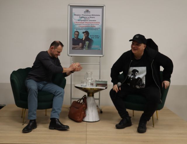 Na zdjęciu znajduje się dwóch mężczyzn siedzących na scenie, mężczyzna po lewo klaszcze. Między mężczyznami stoi stolik, na którym leży woda oraz książki, a za nim stoi plakat promujący spotkanie.