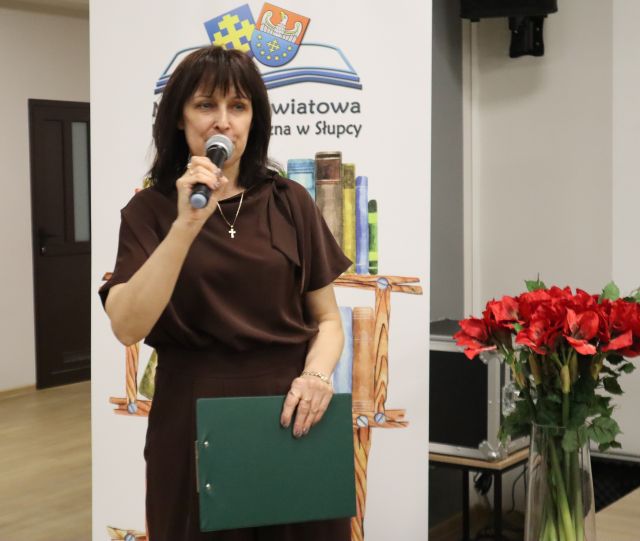 Na zdjęciu znajduje się kobieta od pasa w górę, w jednej ręce trzyma mikrofon, a w drugiej zieloną podkładkę. Za kobietą znajduje się baner z książkami i logiem Biblioteki, po prawej stoją czerwone sztuczne kwiaty.