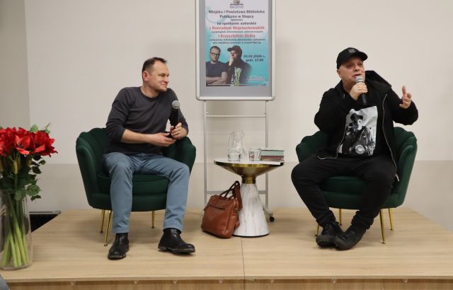 Na zdjęciu znajduje się dwóch mężczyzn siedzących na scenie z mikrofonami w ręku, mężczyzna po prawej gestykuluje. Między nimi stoi stolik z wodą oraz książkami, a za nim plakat promujący spotkanie. Po lewej stronie na scenie stoją czerwone kwiaty.