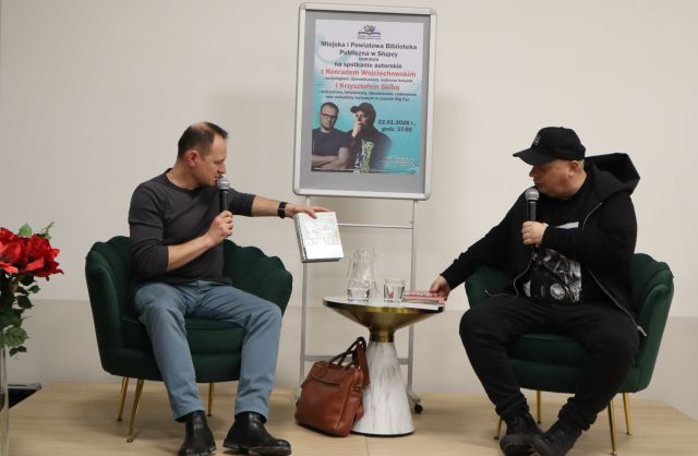 Na zdjęciu znajduje się dwóch mężczyzn na scenie, z mikrofonami. Mężczyzna po lewej sięga książkę ze stojącego pomiędzy nimi stolika, a mężczyzna po prawej trzyma rękę na stoliku. Za mężczyznami znajduje się plakat promujący spotkanie.