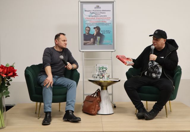 Na zdjęciu znajduje się dwóch mężczyzn na siedzących na scenie, mężczyzna po prawej trzyma w ręce książkę. Pomiędzy nimi stoi stolik z wodą i książkami i torba oparta o stolik. Za mężczyznami znajduje się plakat promujący spotkanie.