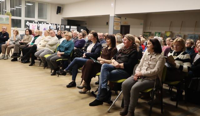 Na zdjęciu znajduje się widownia ma krzesłach, skierowana w stronę sceny. Spotkanie odbywa się na holu, w tle znajdują się okna.