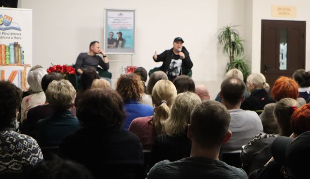 Na zdjęciu znajduje się widownia skierowana w stronę sceny, na której znajduje się Konrad Wojciechowski oraz Krzysztof Skiba, za mężczyznami znajduje się plakat promujący spotkanie, a po lewej stronie przy scenie baner z książkami i logiem Biblioteki.