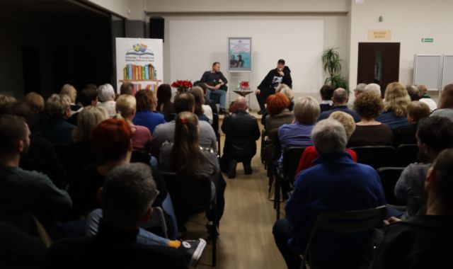 Na zdjęciu znajdują się dwa rzędy widowni i mężczyzna klęczący pomiędzy nimi i robiący zdjęcie mężczyzną na scenie, za któymi stoi plakat promujący spotkanie. Po lewej stronie przy scenie stoi baner z książkami i logiem Biblioteki.