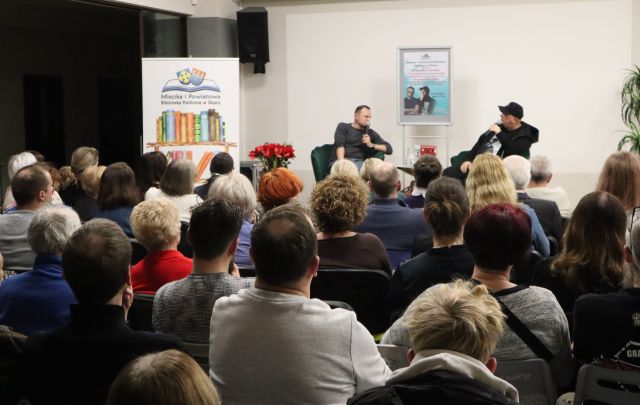 Na zdjęciu znajduje się widownia, tyłem skierowana w stronę sceny, na której siedzi dwóch mężczyzn, za nimi stoi plakat promujący spotkanie. Po lewej stronie na scenie stoją czerwone kwiaty, a obok przy scenie baner z książkami i logiem Biblioteki.