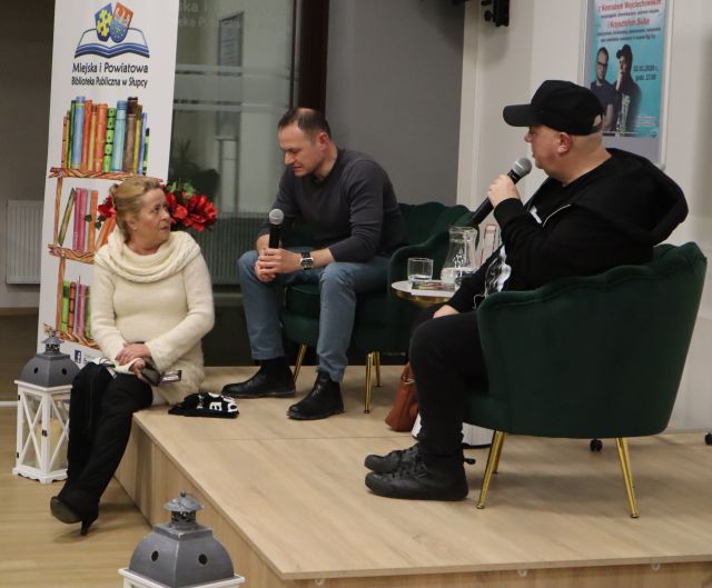 Na zdjęciu znajduje się dwóch mężczyzn siedzących na scenie z mikrofonami oraz jedna kobieta, za którą znajduje się baner z książkami i logiem Biblioteki. Zdjęcie zostało wykonane od boku.