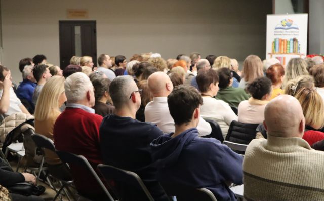 Na zdjęciu znajdują się ludzie na widowni skierowani w stronę sceny. W tle znajduje się baner z książkami i logiem Biblioteki. Zdjęcie zostało wykonane od tyłu.