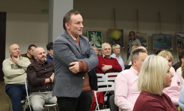 Na zdjęciu znajduje się stojący mężczyzna ze skrzyżowanymi rękoma wśród siedzącej widowni. W tle znajdują się obrazy.