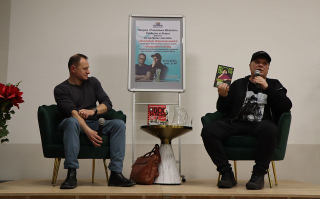 Na zdjęciu znajduje się dwóch mężczyzn na scenie, mężczyzna po prawej trzyma i pokazuje książkę. Między mężczyznami znajduje się stolik i wodą i książkami, a za nim plakat promujący spotkanie.