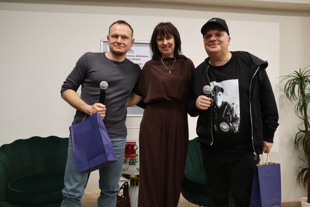 Na zdjęciu znajduje się dwóch mężczyzn i kobieta między nimi, stoją do zdjęcia. Mężczyźni trzymają w ręce mikrofony i granatowe papierowe torby. W tle znajdują się fotele oraz plakat.