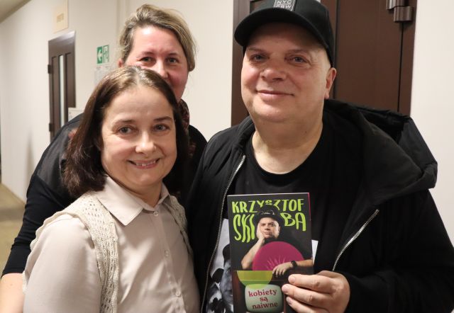 Na zdjęciu znajdują się dwie kobiety, które stoją do zdjęcia z Krzysztofem Skibą, który trzyma w ręce książkę.