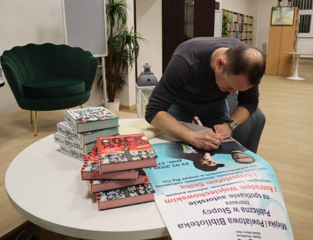 Na zdjęciu znajduje się Konrad Wojciechowski nachylony nad stołem i podpisujący plakat promujący spotkanie, obok leżą dwie kupki książek. A w tle znajduje się scena i fotelami.