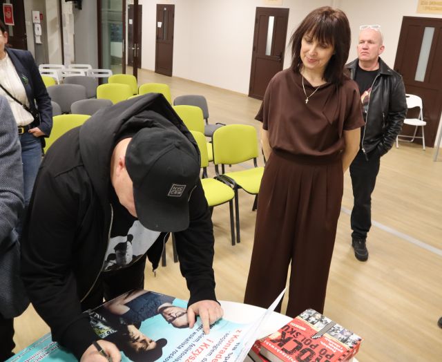 Na zdjęciu znajduje się Krzysztof Skiba nachylony nad stołem i podpisujący plakat, po prawej stronie stoi kobieta i mężczyzna za nią. W tle znajdują się krzesła.