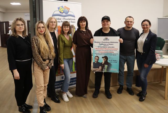 Na zdjęciu znajduje się sześć kobiet stojących do zdjęcia z Konradem Wojciechowskim oraz Krzysztofem Skibą, który trzyma plakat promujący spotkanie. W tle znajduje się baner z logiem Biblioteki.
