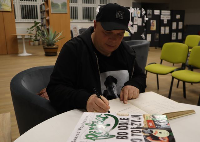 Na zdjęciu znajduje się Krzysztof Skiba przy stole i wpisuje się do Kroniki, na stole znajdują się również książki, a w tle krzesła oraz półki.