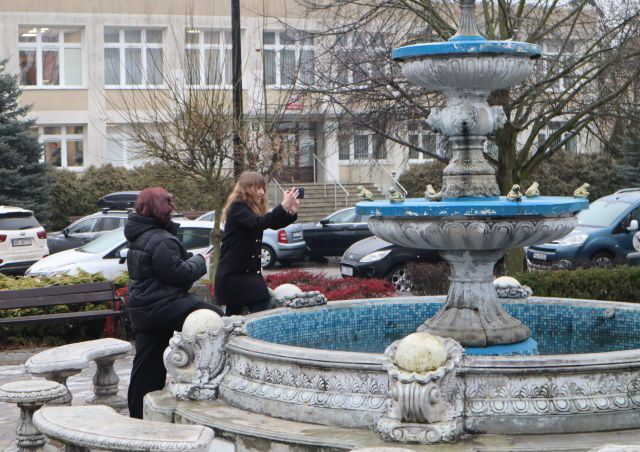 Na zdjęciu znajdują się dwie nastolatki przy fontannie, jedna robi zdjęcie. W tle znajdują się drzewa, stojące auta i budynek.