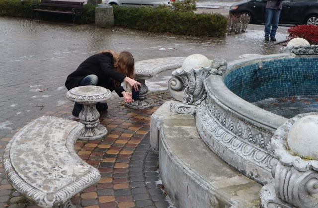 Na zdjęciu znajduje się kucająca nastolatka i robiąca zdjęcie fontannie przed sobą. Dookoła fontanny stoją ławki. W tle znajdują się krzaki.