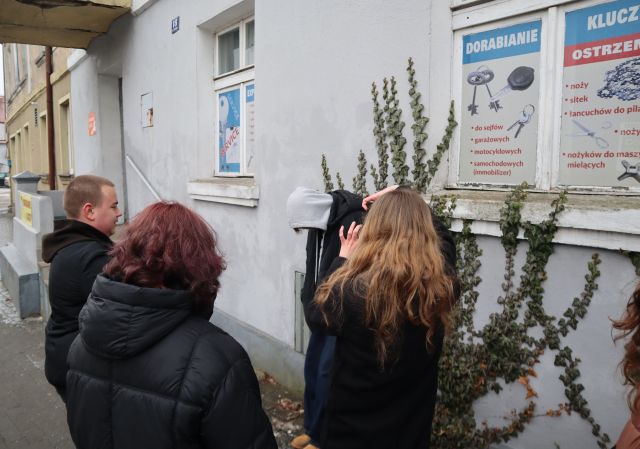 Na zdjęciu znajduje się trójka osób. Jedna dziewczyna robi zdjęcie osobie stojącej przy budynku, na którym rośnie winorośl.