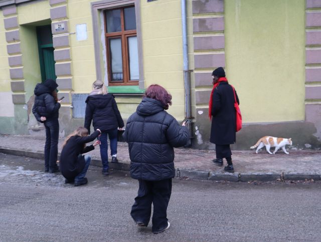 Na zdjęciu znajduje się pięć dziewczyn, jedna z nich klęka i robi zdjęcie przechodzącemu kotu. Przed dziewczynami znajduje się stary zielony budynek.