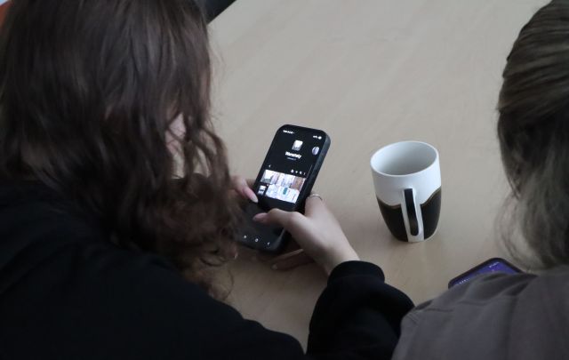 Na zdjęciu znajdują się dwie dziewczyny przerabiające zdjęcia na telefonie, przed nimi na stoliku stoi kubek. Zdjęcie zostało wykonane od tyłu z góry.