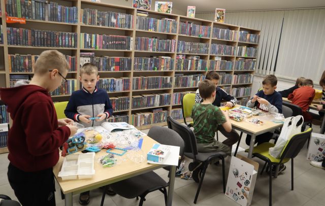 Na zdjęciu znajdują się trzy stoliki, przy pierwszym stoi dwóch chłopców i układają LEGO, a przy drugim i trzecim trzech chłopców. Na stolikach znajdują się klocki LEGO, a koło stolików kartony. W tle znajdują się półki z książkami.