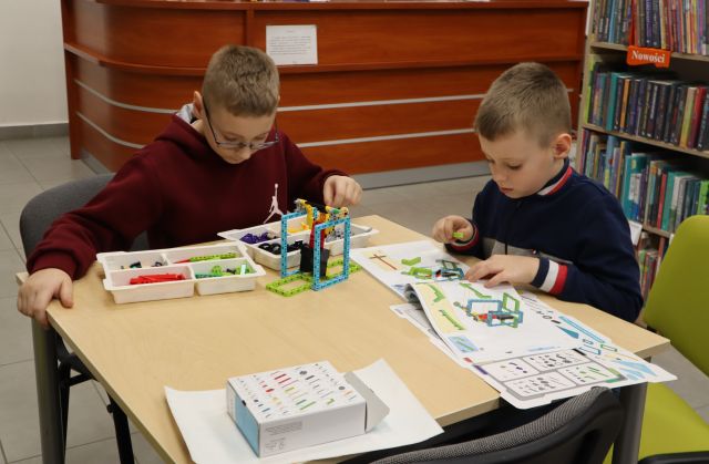 Na zdjęciu znajduje się dwóch chłopców przy stoliku i układają LEGO według instrukcji. W tle znajduje się biurko oraz półki z książkami.
