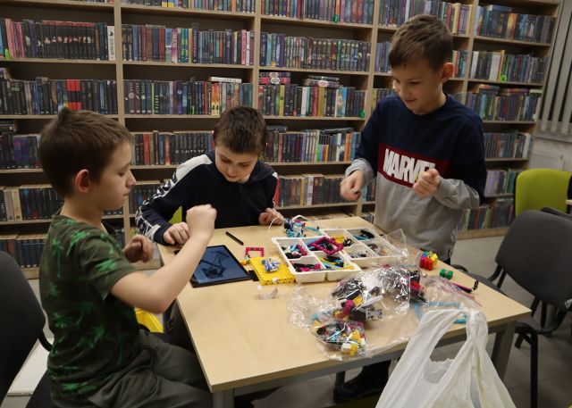 Na zdjęciu znajduje się trzech chłopców, dwóch siedzi a jeden stoi przy stoliku i układają LEGO według instrukcji wyświetlanej na tablecie. Na stoliku leżą klocki, a na wolnym krześle przy stoliku leży plastikowa torba. W tle znajdują się półki z książkami.